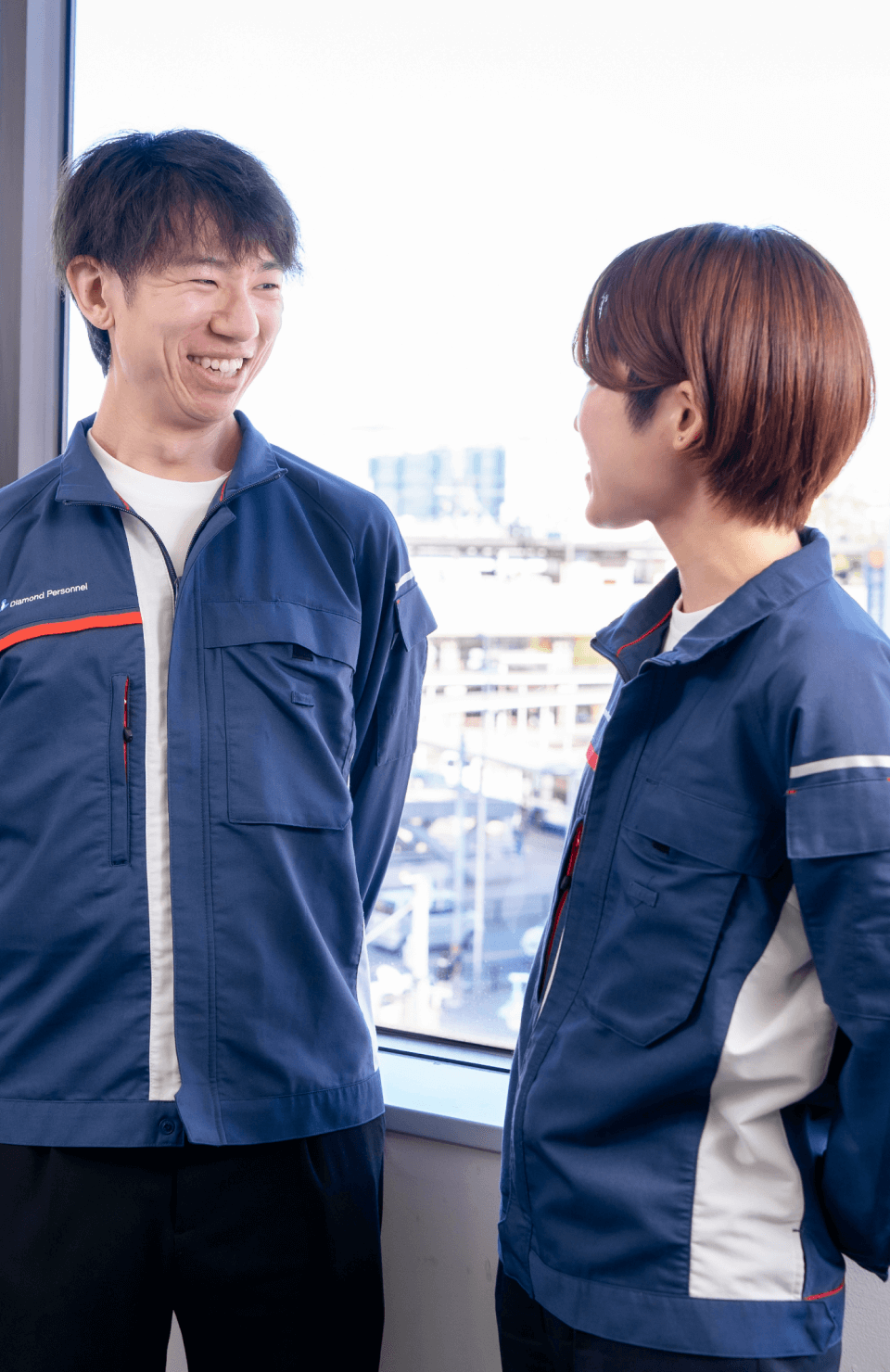 青い作業服を着た男性と女性が窓際で笑顔で会話している