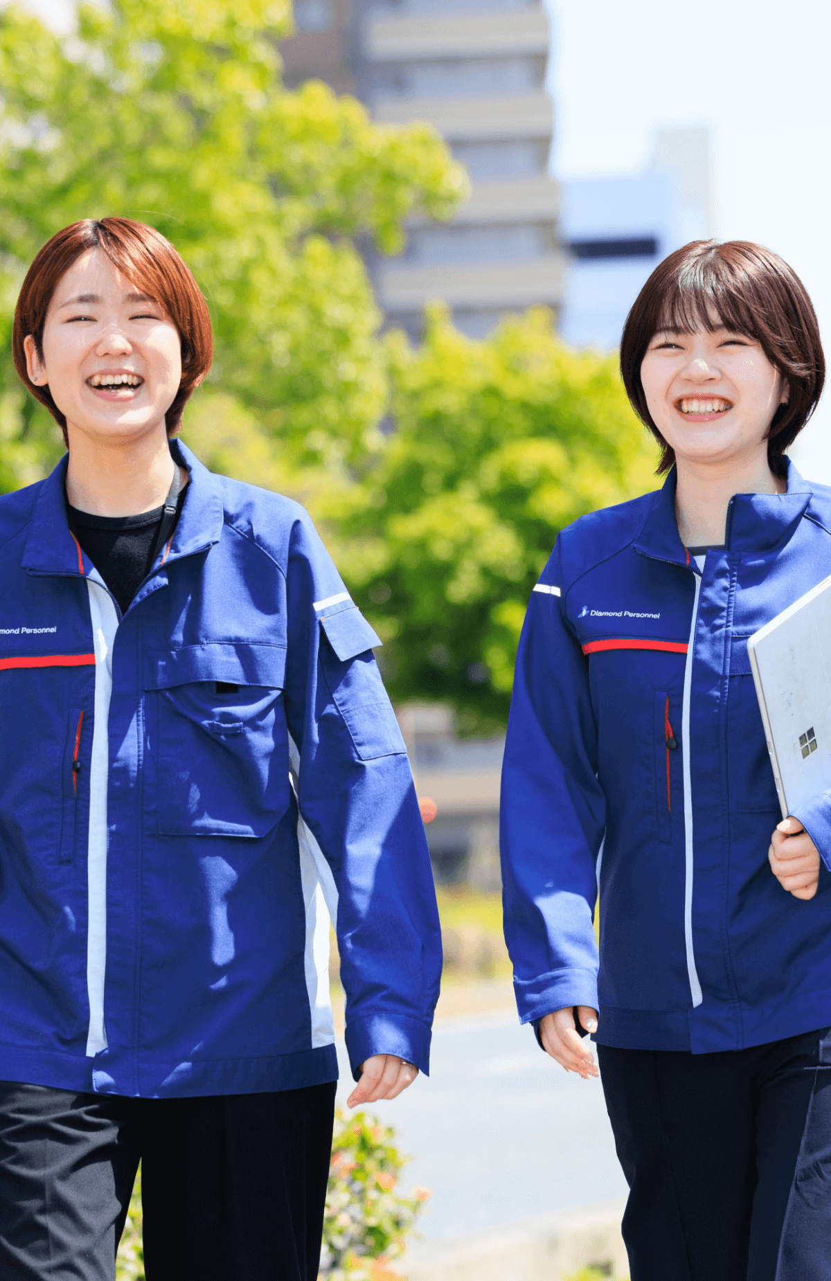 屋外で青い作業服を着た二人の女性が並んで歩きながら笑顔を見せている
