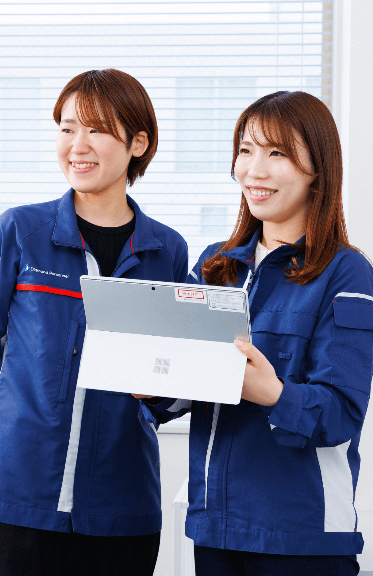 青い作業服を着た二人の女性がタブレット端末を持ちながら微笑んでいる室内の写真