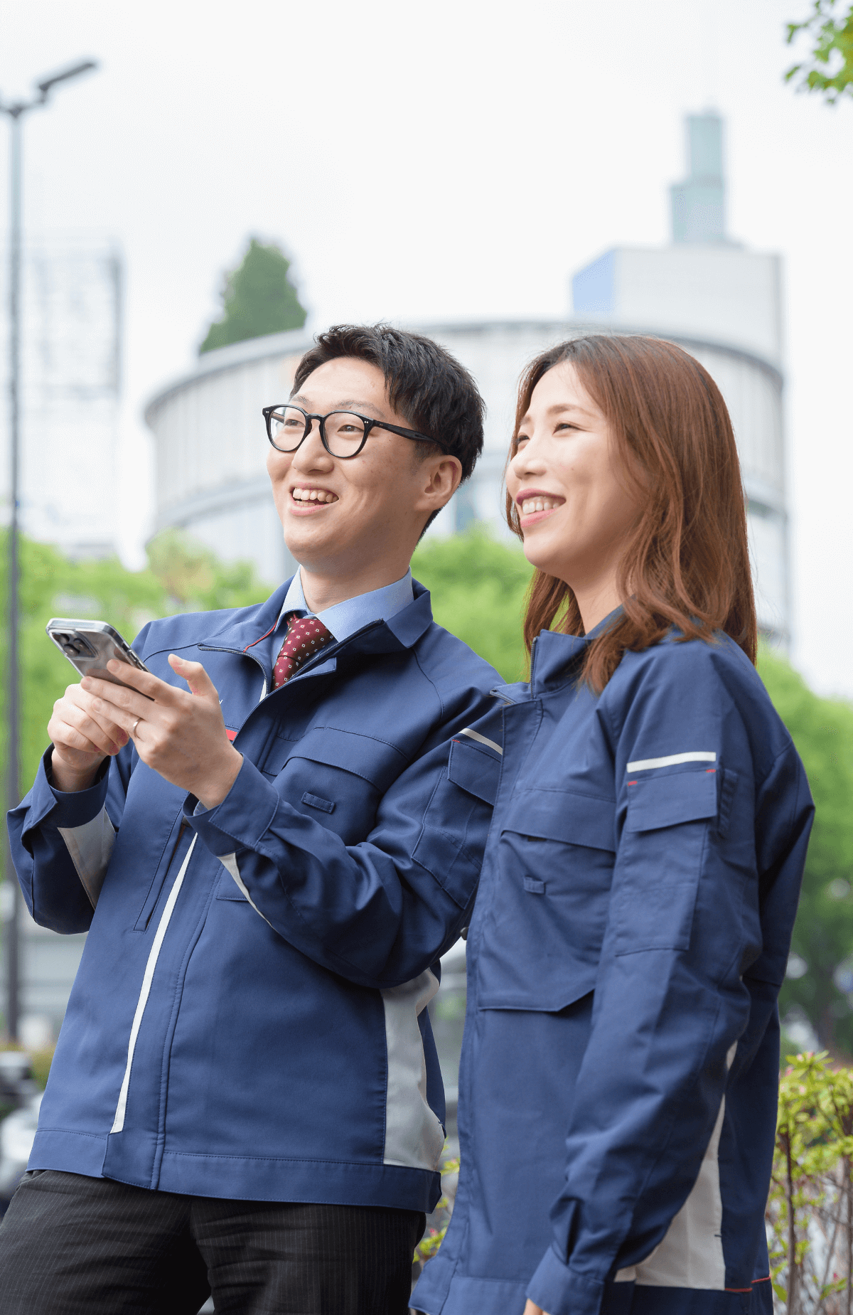 屋外で青い作業服を着た男性と女性が並び、男性がスマートフォンを手に持ちながら笑顔で話している